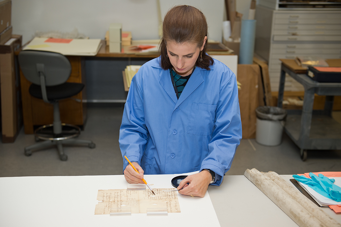 Women in blue lab coat stands at table. She takes a small paint brush and tools to treat a document.