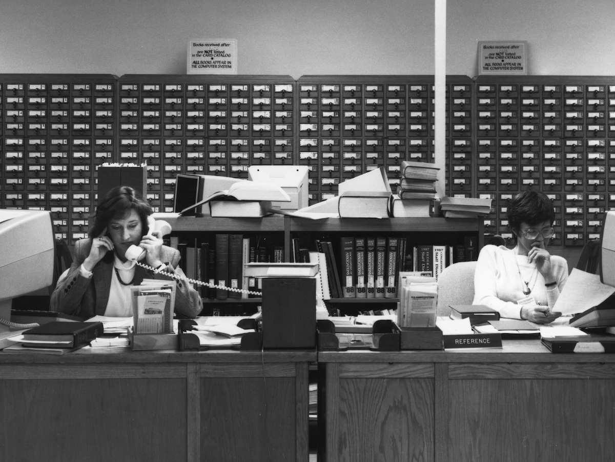 Two women sit at a desk, one of the phone and the other reading a piece of paper. 