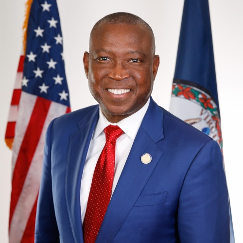 African American man wearing a blue jacket and red tie in front of the American and Virginia flags