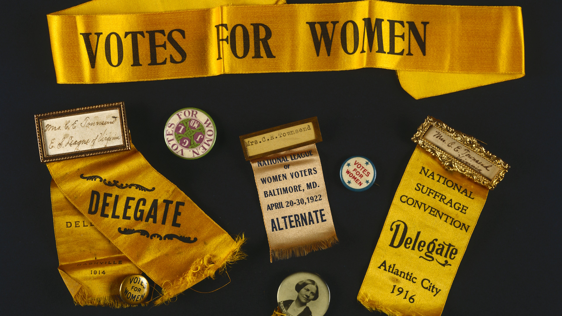 yellow sash and two pinback buttons reading Votes for Women and suffrage convention badges with yellow ribbons