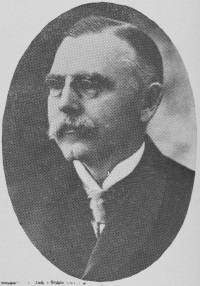 Headshot of a white man with a mustache wearing a suit and tie.