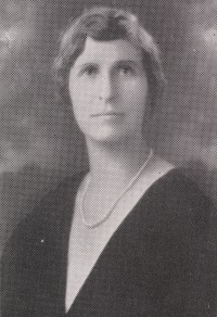 Headshot of a white woman wearing pearls and a black dress.