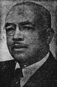 Newspaper photograph of an African American man with a mustache wearing glasses and a suit and tie.