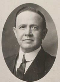 A white man wearing a mid-20th century suit and tie.