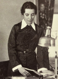 White woman in early 20th century attire standing at a desk looking at a book.