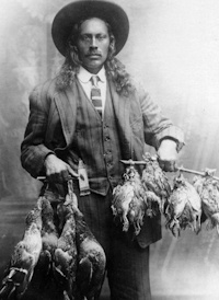 Native American man wearing early 20th-century attire holding two braces of dead fowl in his hands.