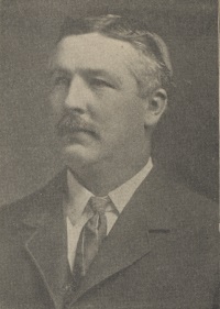 Headshot of a white man with a mustache wearing a mid-20th century suit and tie.
