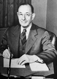 A white man wearing glasses and a mid-20th century suit and tie seated at a desk with a pen in his hand.