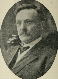 White man with a mustache wearing glasses and a suit and tie.