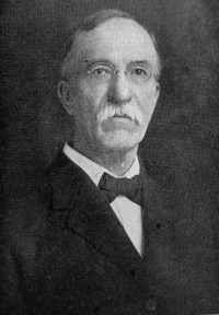 Headshot of a white man with a mustache and wearing glasses and a suit with a bowtie.