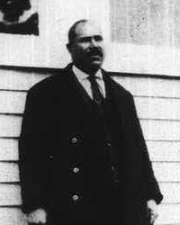 African American man with a mustache wearing early 20th century attire standing in front of a clapboard building.