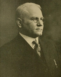 A white man with pinze nez wearing an early 20th century suit and tie.