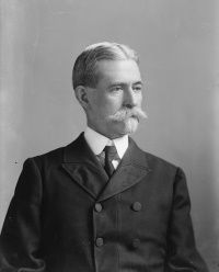 A white man with a mustache and goatee wearing an early 20th century suit and tie.
