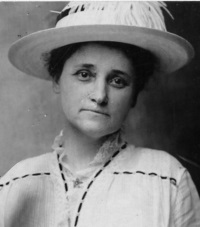 White woman wearing an early 20th century hat and white dress.