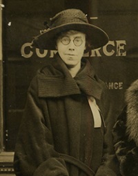 White woman with glasses wearing early 20th century hat and coat.