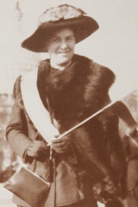 White woman wearing a 1910s era hat and fur stole carrying a small woman suffrage flag.