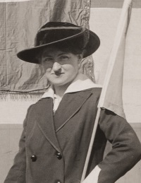 White woman wearing a 1910s era hat and coat outdoors.