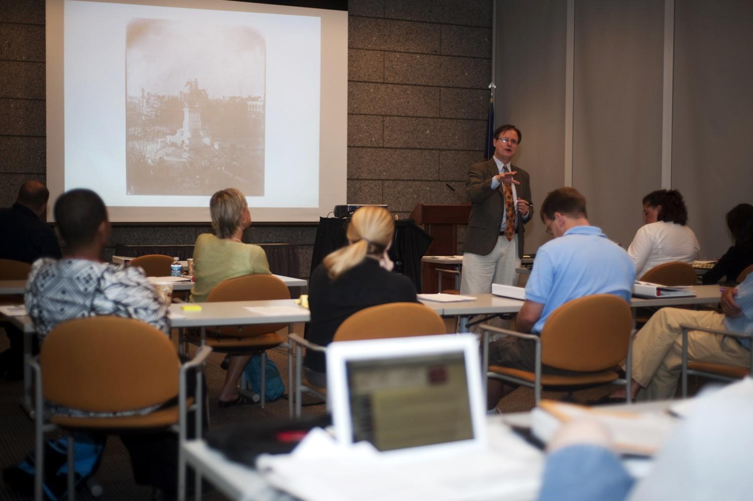 2023 Brown Teacher Institute five people seated a tables facing a man speaking at the front of the room