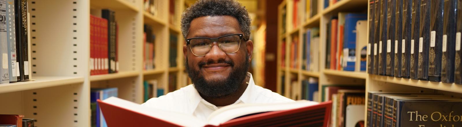 Bilal holding a book. This image shows a man holding a book open while standing in between rows of book shelves.