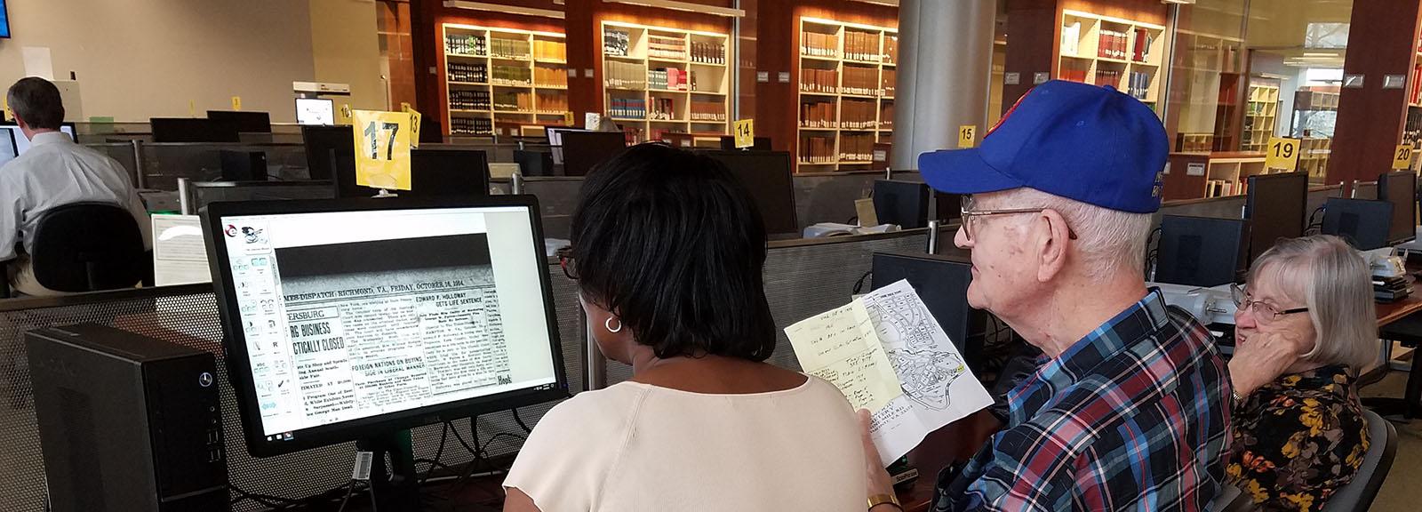Library of Virginia patrons viewing newspaper microfilm in the Library's reading room. Two women and a man in a blue cap looking at a computer with an image from a newspaper on it.