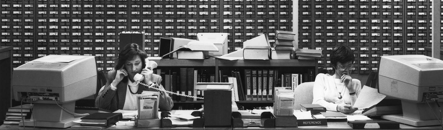 Mary Sine Clark and Sarah Huggins, Library of Virginia reference. Two women sitting at a long desk, with computer terminals next to them. One is on the phone, while the other reads a piece of paper. Behind them is a large card catalog.