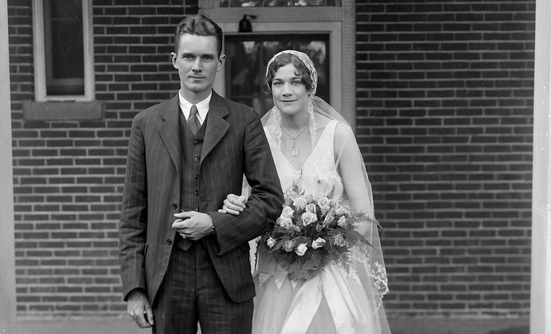 Unidentified group portrait. M03504. Michael Miley Collection. Library of Virginia. A wedding photograph featuring a man in the three-piece suit and a bride in sleeveless white dress to below her knees. She wears white stockings and shoes and a veil. She carries a bouquet of light colored roses.
