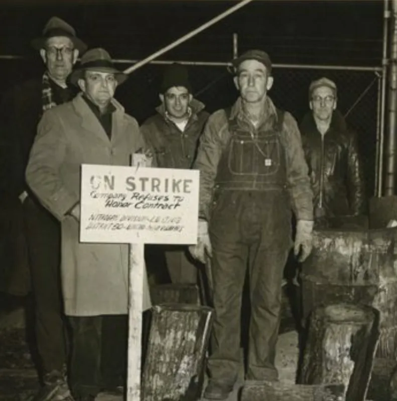 Five men standing behind a small sign that says "On Strike Company Refuses to Honor Contract." Two men are wearing top coats and fedoras, while the other three are wearing work clothes, such as overalls, leather jackets and caps. 