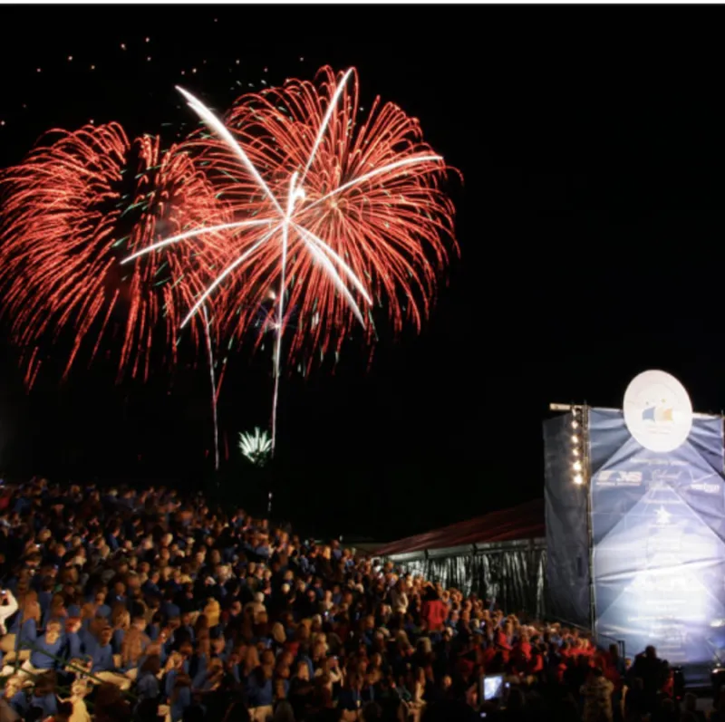 Image shows red and white fireworks lighting the night sky above a large crowd.