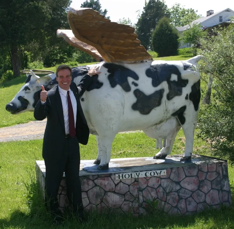 Virginia Governor Mark Warner stands next to a large statue of a black and white cow with wings. The cow stands on a pedestal that says "Holy Cow." Warner is wearing a dark suit with a white dress shirt and red tie. He is smiling and is "giving a thumbs up."