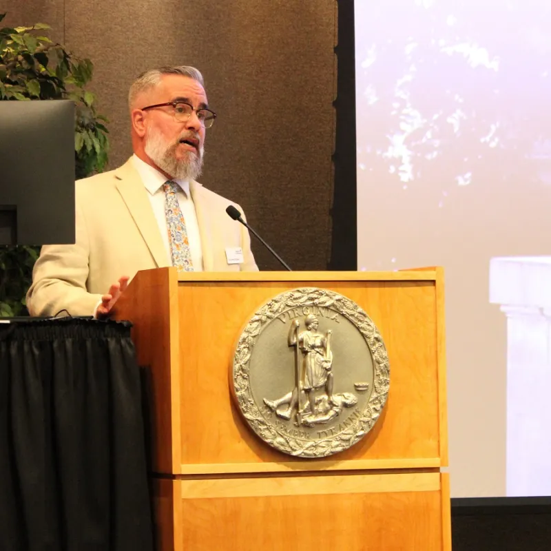 man in tan suit speaks into a microphone while presenting at a podium